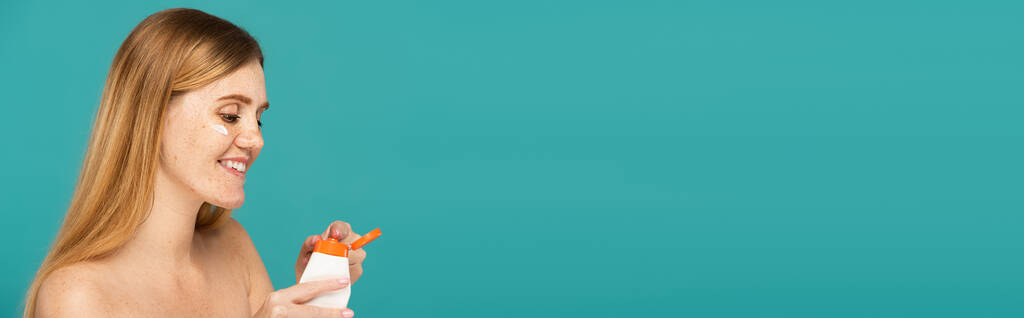 veselá zrzka žena s pihami drží trubice s hydratační krém izolované na tyrkysové, banner - Fotografie, Obrázek
