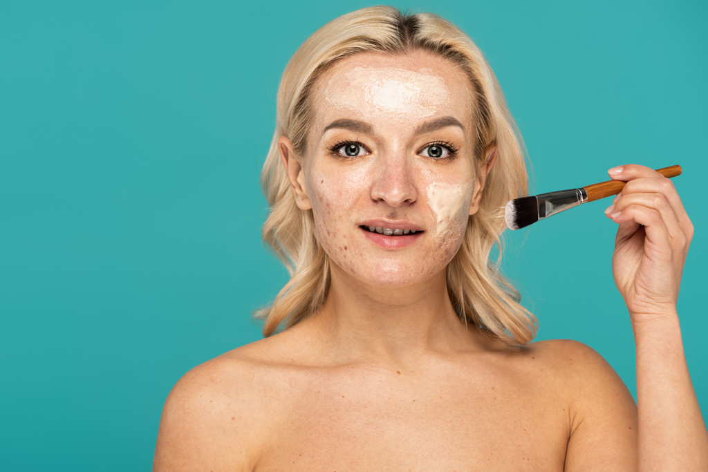 Blondine mit Akne hält Kosmetikpinsel in der Hand und trägt Tonmaske isoliert auf Türkis auf  - Foto, Bild