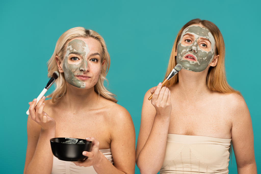 mujeres rubias y pelirrojas aplicando máscara de arcilla en las caras con cepillos cosméticos aislados en turquesa - Foto, imagen