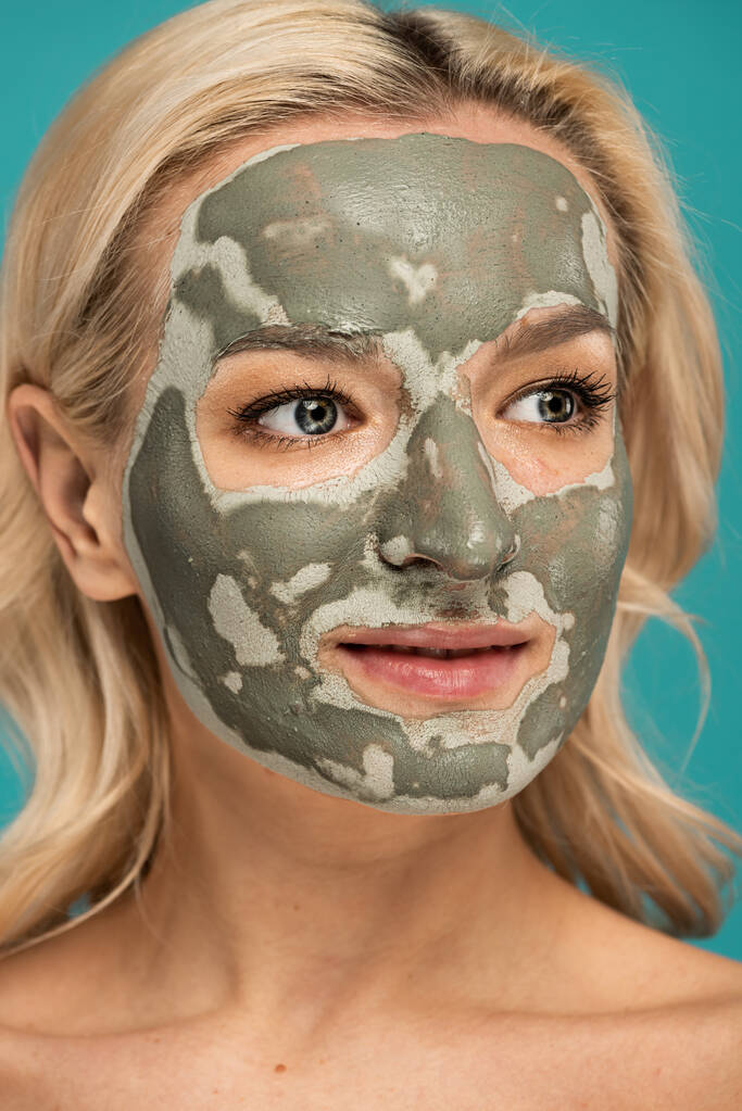 portrait de femme blonde avec masque d'argile sur le visage regardant loin isolé sur turquoise - Photo, image