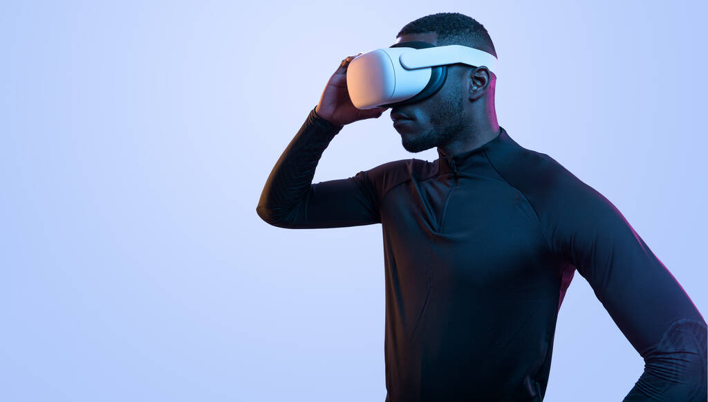 Zijaanzicht van ernstige bebaarde Afro-Amerikaanse man in zwarte coltrui aanraken virtual reality googles terwijl staan op blauwe achtergrond - Foto, afbeelding