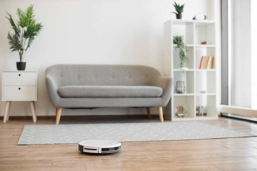 Advanced robotic vacuum cleaner hoovering airy living room in cozy apartment during sunny day. Efficient household appliance keeping hard floor and soft carpet tidy via remote control or phone app. - Photo, Image