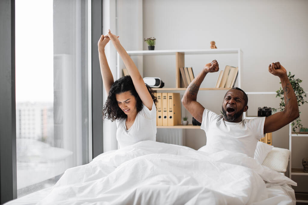 Szerető afro-amerikai pár alkalmi öltözékben nyújtják ki a végtagjaikat az éjszakai alvás után, miközben még mindig a fényes hálószoba ágyában fekszenek. Pihentetett család férfi és nő új napsütéses nap a modern lakásban. - Fotó, kép