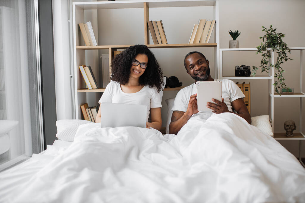 Happy multiracial married couple dealing with smart gadgets while relaxing under cozy duvet in airy bedroom of modern apartment. Cheerful family man and woman uploading content to social media. - Photo, Image