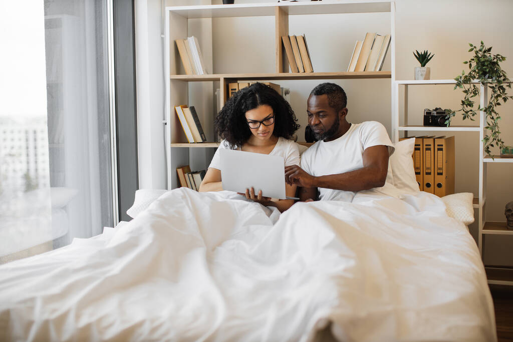 美しい若い女性がベッドに座って、彼女の夫がアドバイスで彼女を支援しながら、現代のラップトップを使用してテキストメッセージを送信します。家庭で休息技術生活のバランスを達成する夫婦を愛し. - 写真・画像