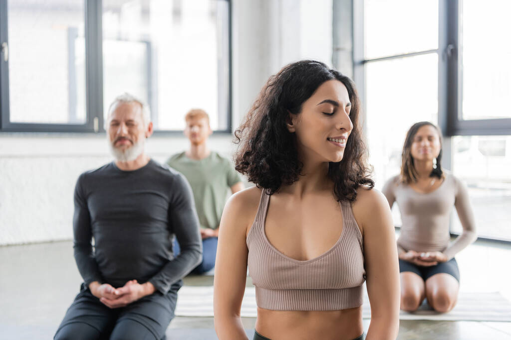 Sorridente donna mediorientale meditare vicino persone offuscate in classe di yoga  - Foto, immagini