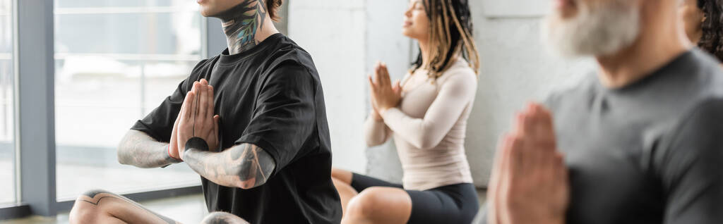 Hombre tatuado haciendo anjali mudra cerca de grupo interracial en clase de yoga, pancarta  - Foto, Imagen