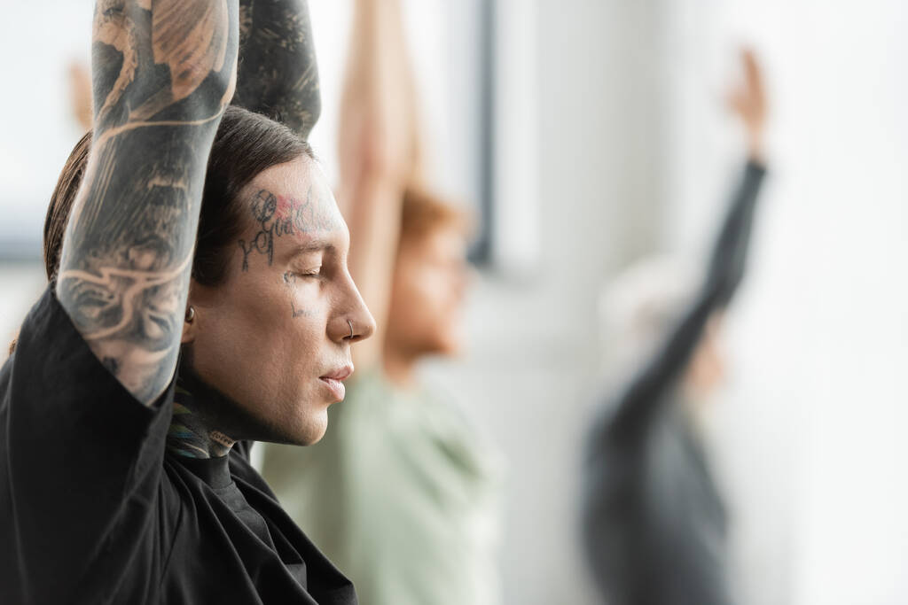 Вид сбоку на молодого человека с татуировками, медитирующего на занятиях йогой  - Фото, изображение