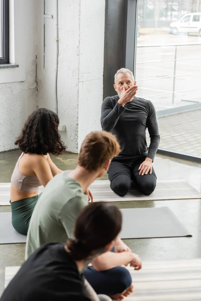 Rijpere coach toont neusgat ademhaling naar wazig groep in yoga studio  - Foto, afbeelding