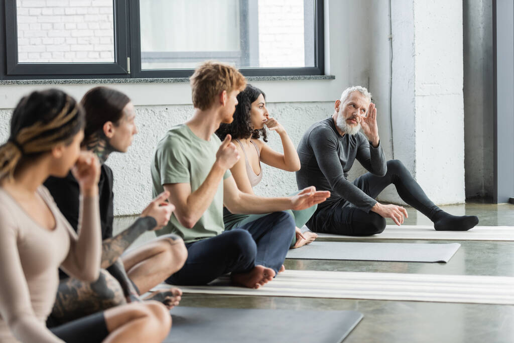Middelbare leeftijd coach uitleggen neusgat ademhaling aan multi-etnische groep in yoga klasse  - Foto, afbeelding