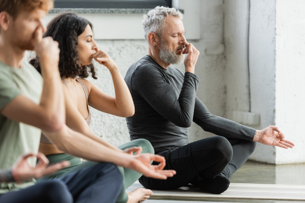 Uomo maturo che pratica la respirazione narice e mudra gyan vicino a persone interrazziale in studio di yoga  - Foto, immagini