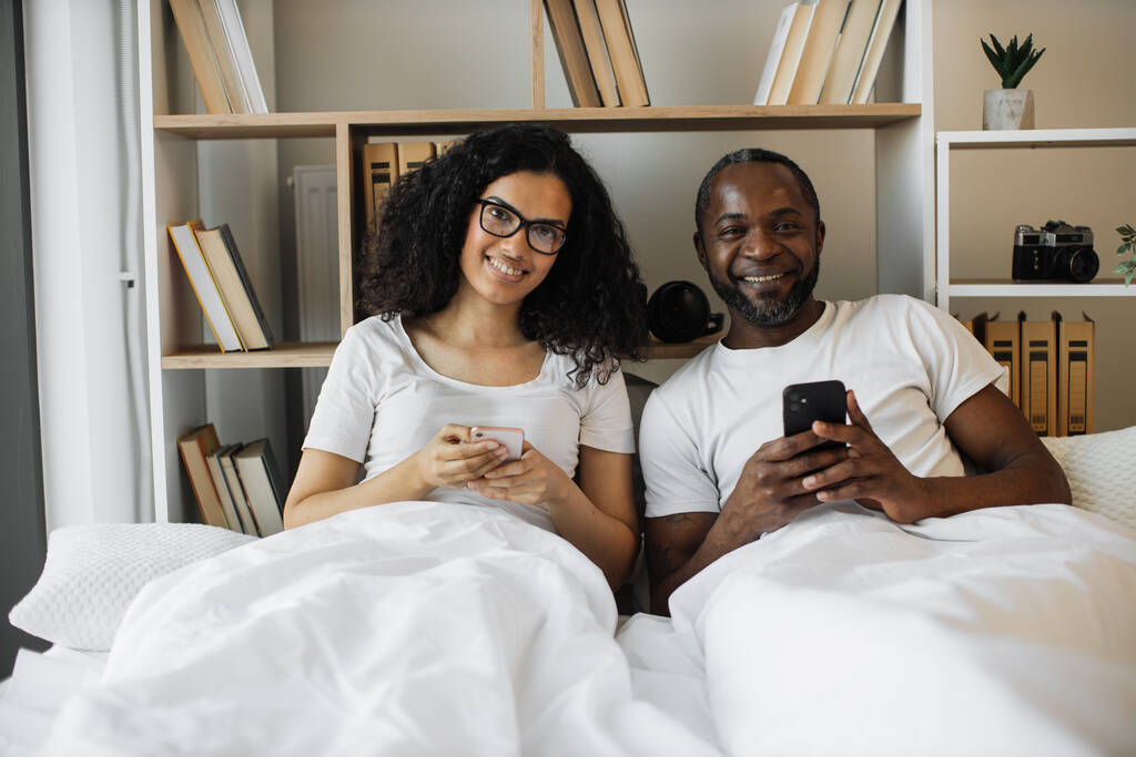Feliz pareja afroamericana sosteniendo teléfonos celulares en las manos mientras descansa en una cama suave toda la mañana en el interior. Atractivo hombre y mujer navegando por Internet utilizando la aplicación móvil en dispositivos digitales. - Foto, imagen
