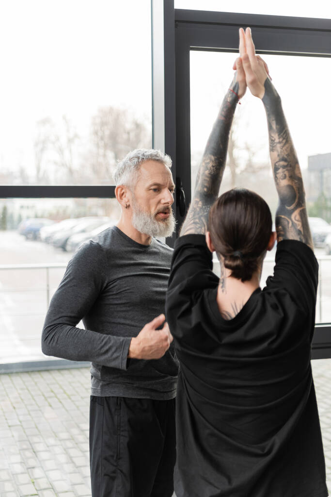 Coach mature parlant à un homme tatoué faisant Crescent Lunge asana dans un studio de yoga  - Photo, image