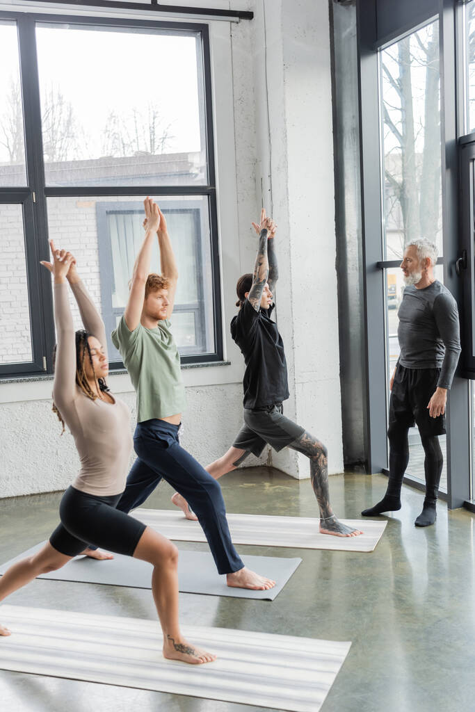 Międzyrasowe osoby stojące w Crescent Lunge asana w pobliżu dojrzałego trenera na zajęciach jogi  - Zdjęcie, obraz