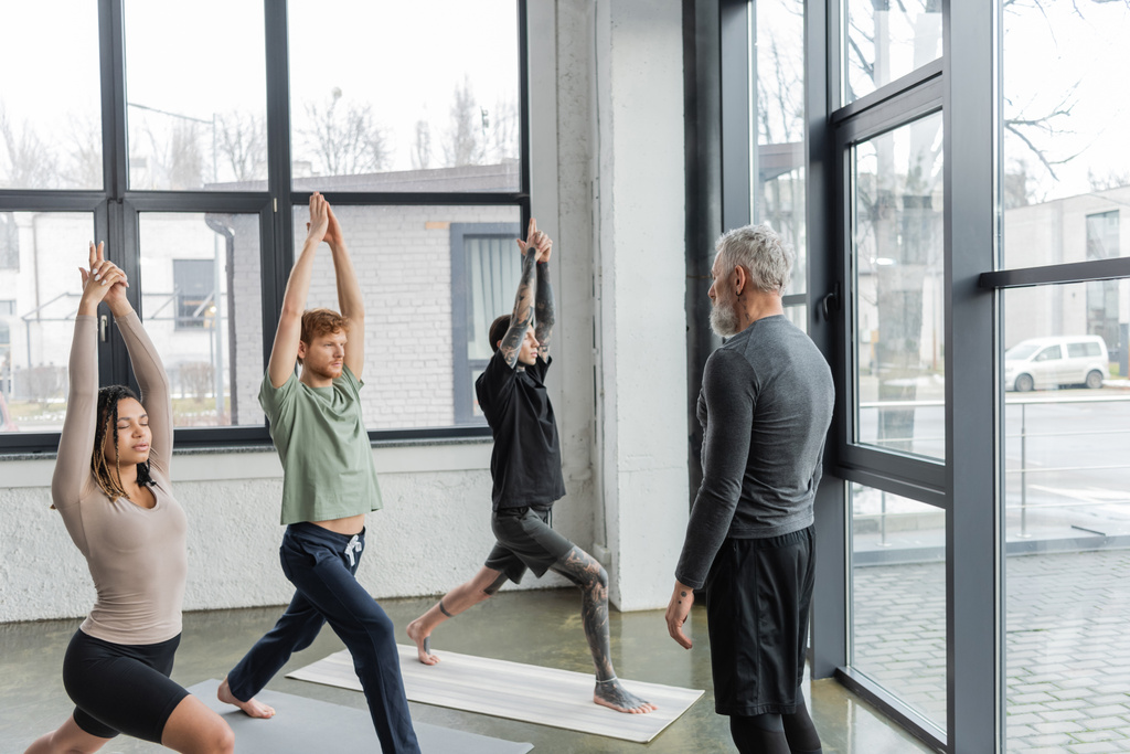 Trener w średnim wieku stojący w pobliżu wielonarodowej grupy ćwiczącej pozycję Crescent Lunge w studiu jogi  - Zdjęcie, obraz