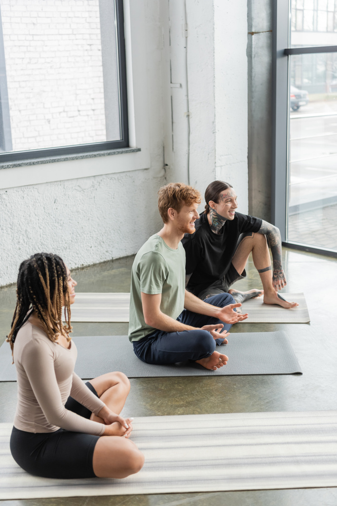 Junge lächelnde multiethnische Menschen unterhalten sich im Yoga-Kurs auf Matten  - Foto, Bild