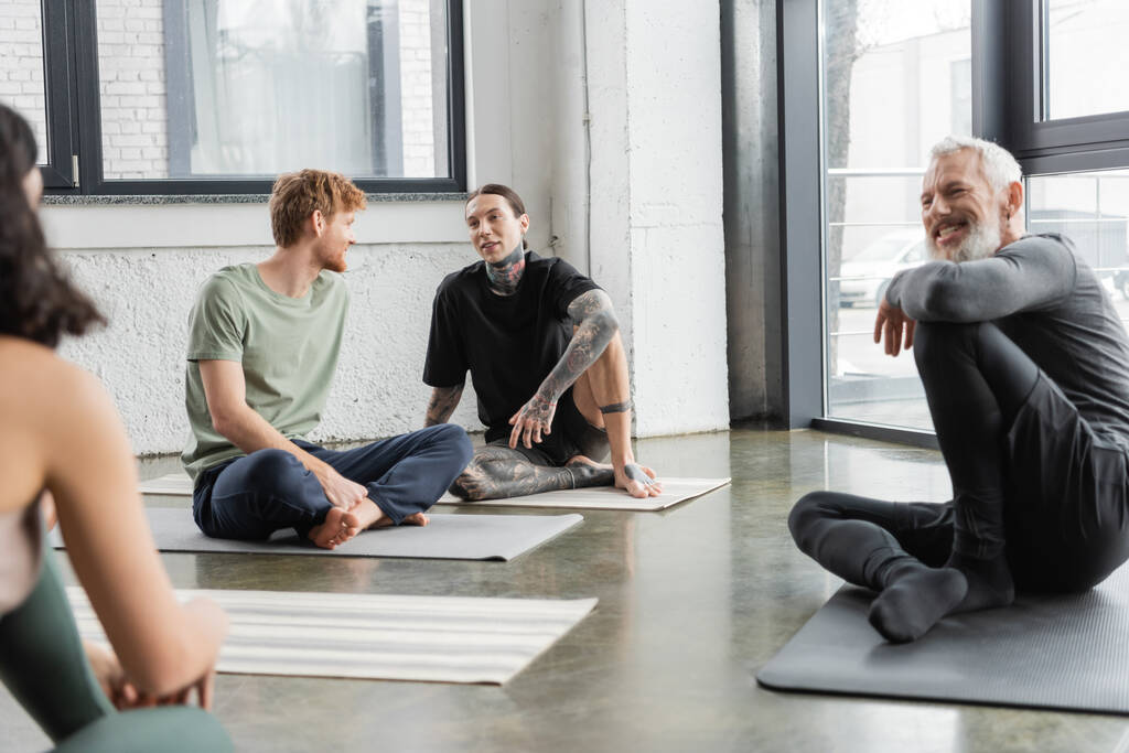 Giovani uomini che parlano mentre seduti su stuoie in classe di yoga  - Foto, immagini