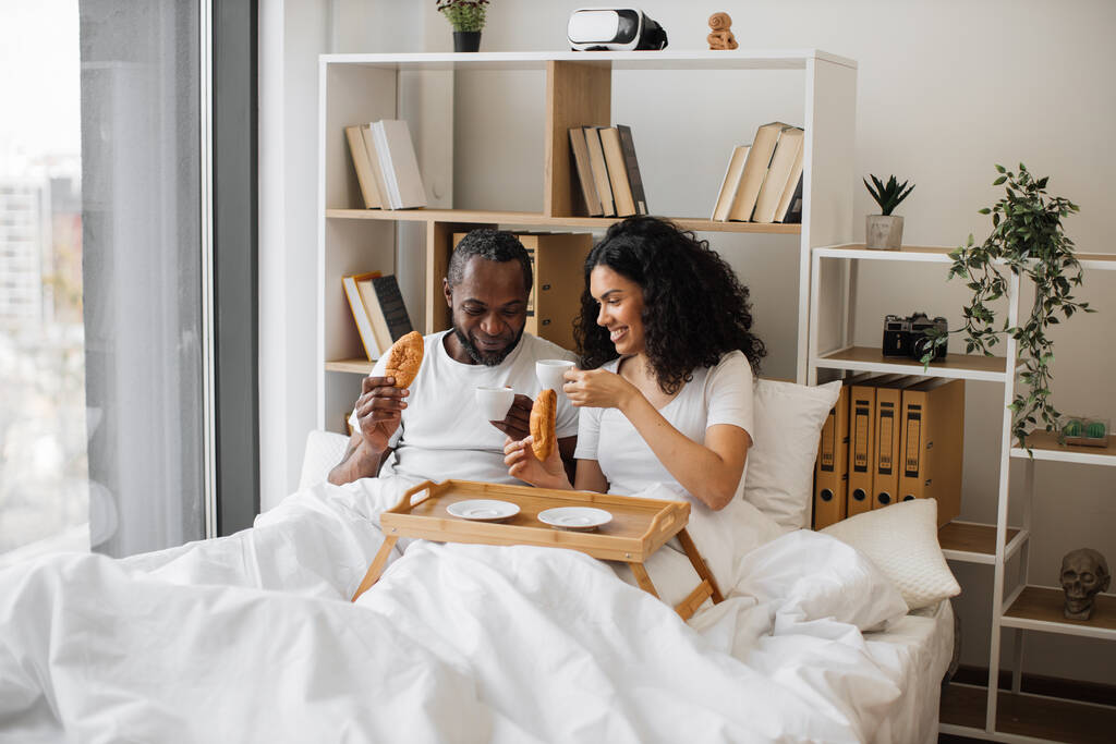 Африканський американський сім'янин і жінка, які їдять півмісяць і п'ють чорну каву, лежачи на ліжку в сонячний ранок. Радісна подружня пара щодня посилає звістку правдивої любові.. - Фото, зображення