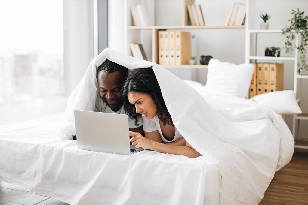 Boldog afro-amerikai házaspár hordozható számítógéppel, miközben a takaró alatt fekszenek otthon, a kényelmes hálószobában. Vidám férj és feleség élvezi időt együtt hétvégén, miközben böngészés online weboldalak. - Fotó, kép