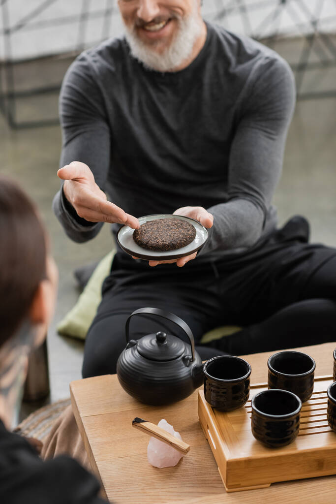bijgesneden blik van gelukkige man van middelbare leeftijd met grijze baard met samengeperste puer thee in yoga studio  - Foto, afbeelding