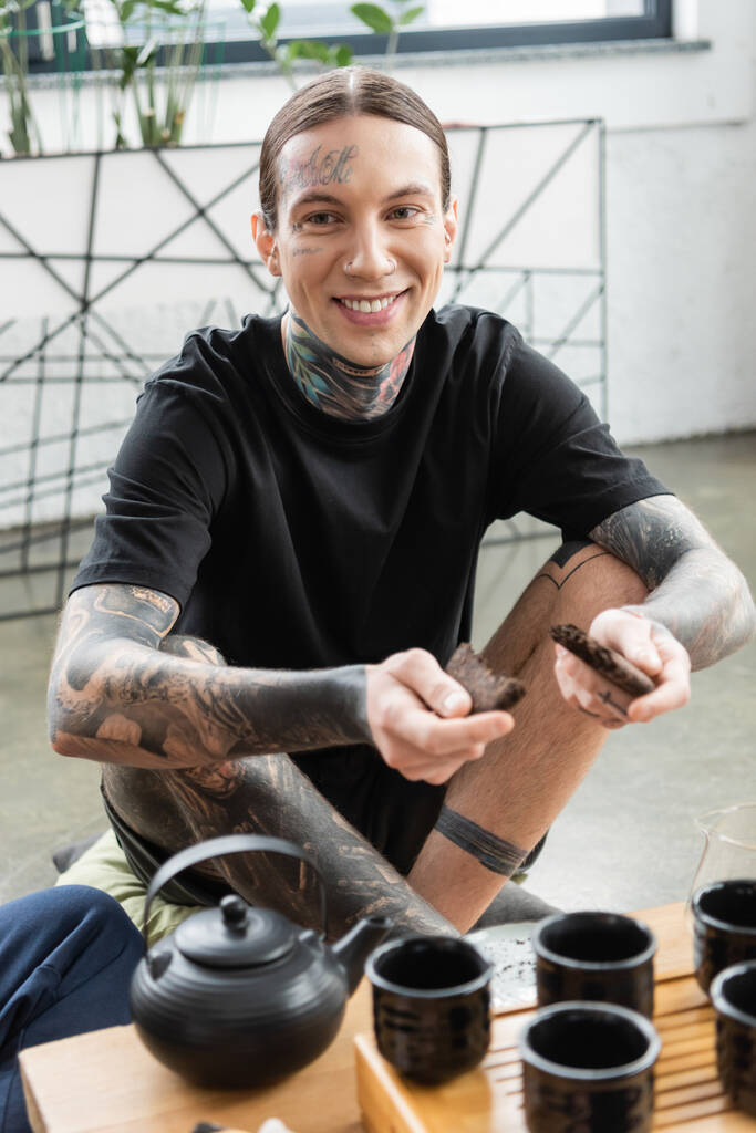 vrolijke jongeman met tatoeages breken samengeperste puer thee in de buurt van traditionele Chinese theepot en bekers  - Foto, afbeelding