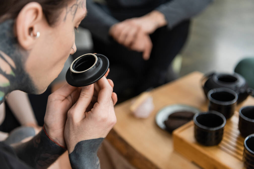 joven tatuado oliendo té puro cerca de tazas chinas en estudio de yoga  - Foto, Imagen