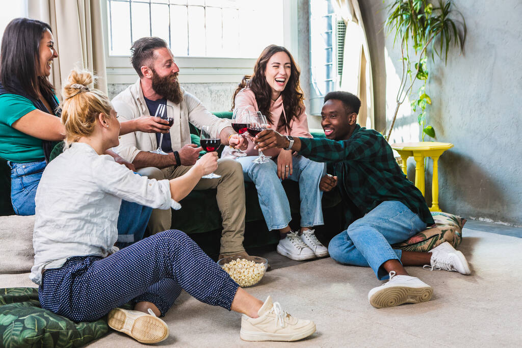 Auf dem Sofa sitzt eine Gruppe von Freunden, die mit Rotwein auf den Aperitif anstoßen. Millennial multiethnische Menschen genießen es, zusammen zu feiern. Lebensstilkonzept mit glücklichen Jungs und Mädchen unterschiedlicher Kultur - Foto, Bild