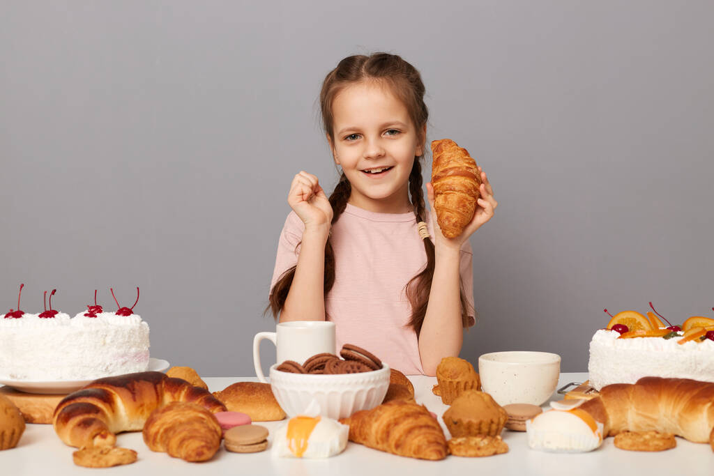 Aranyos, bájos, vidám, copfos nőstény gyermek portréja az asztalnál ülve, szürke háttérrel izolált cukros desszertekkel, croissant-t tartva a kezében, kamerába nézve. - Fotó, kép