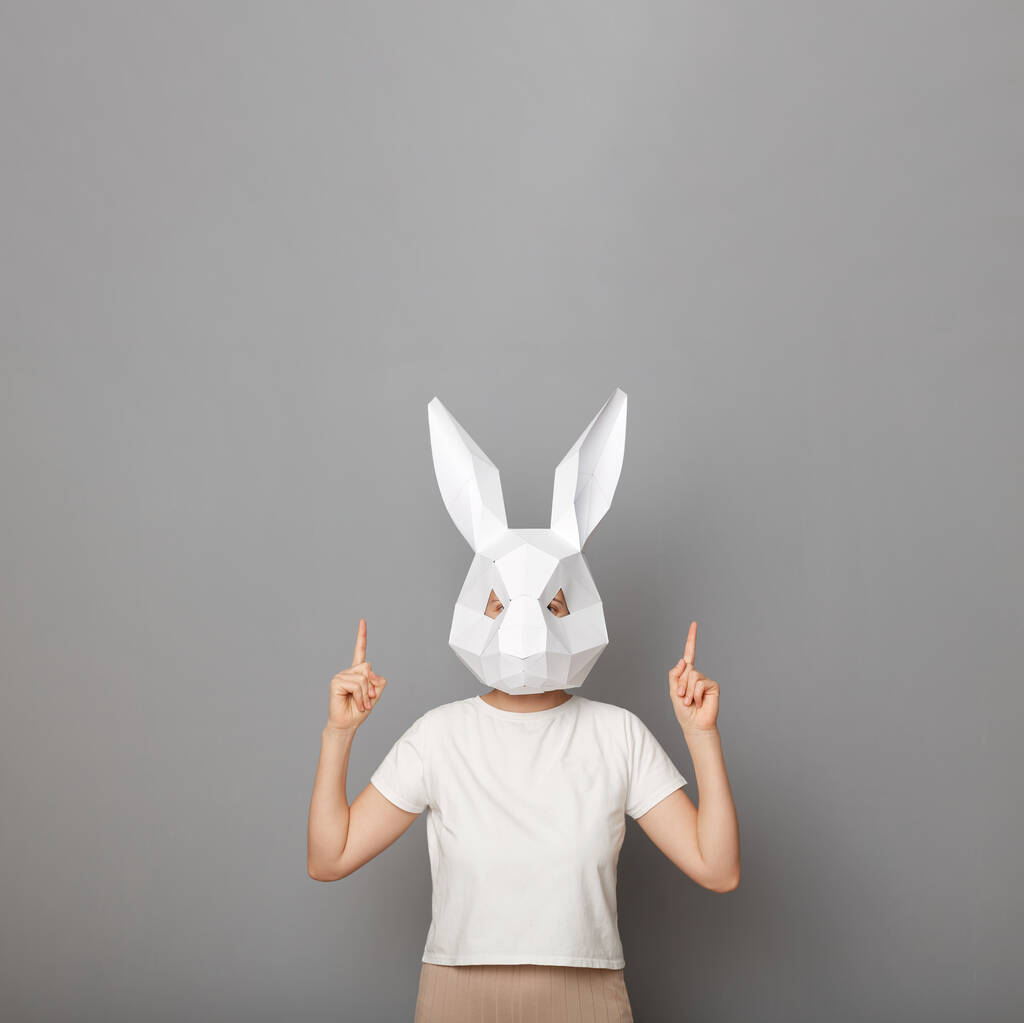 Indoor shot of slim unknown woman wearing white t shirt and paper rabbit mask pointing up at empty space, copy space for advertisement, standing isolated over gray background. - Photo, Image