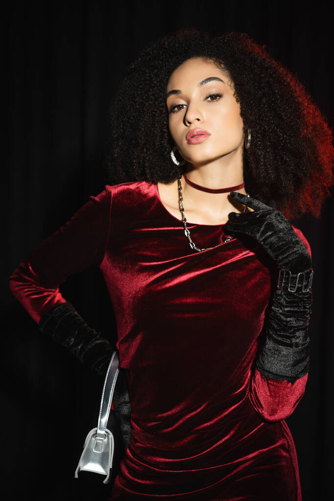 Fashionable african american woman in burgundy dress and gloves posing isolated on black  - Photo, Image