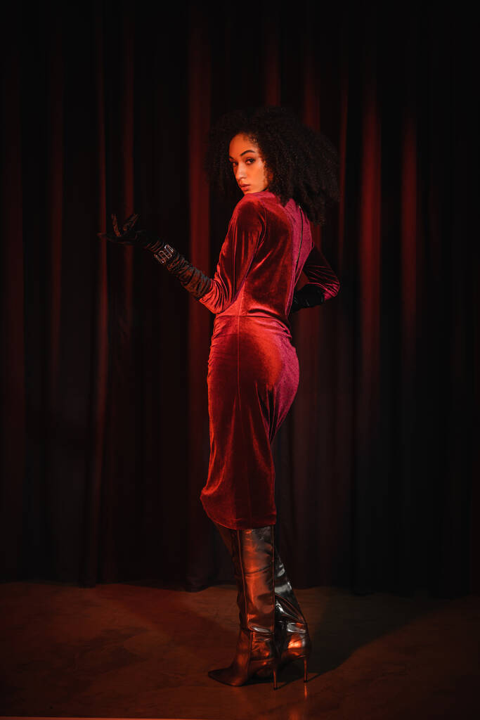 Μοντέρνο αφροαμερικανός γυναίκα σε μπότες γόνατο και φόρεμα κοιτάζοντας κάμερα κοντά στο κόκκινο drapery  - Φωτογραφία, εικόνα