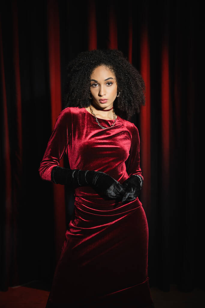 Elegante modelo afroamericano en vestido de terciopelo y guantes mirando a la cámara cerca de cortinas rojas  - Foto, imagen