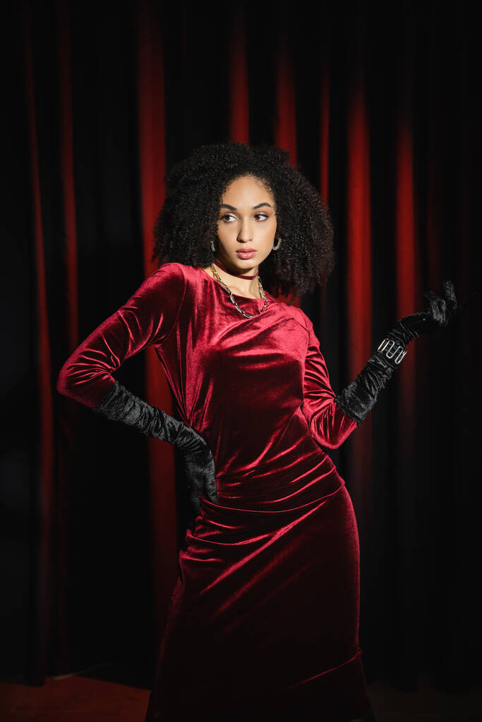 Stilvolle afrikanisch-amerikanische Frau in Handschuhen und Kleid posiert in der Nähe von roten Vorhängen  - Foto, Bild