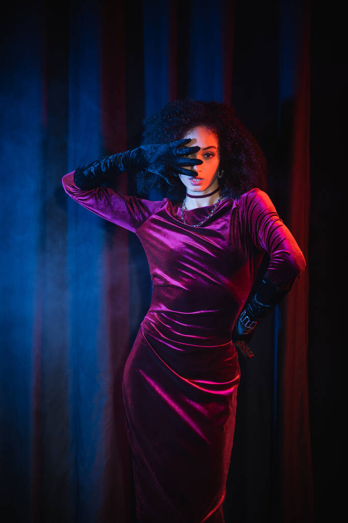 Trendiges afrikanisch-amerikanisches Model in Samtkleid und Handschuhen auf rotem Hintergrund mit blauem Licht - Foto, Bild