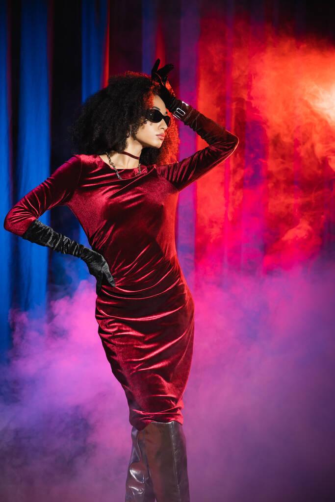 Kırmızı arka planda mavi ışık ve dumanla poz veren kadife elbiseli ve güneş gözlüklü Afro-Amerikan model.  - Fotoğraf, Görsel