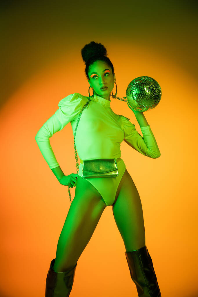 Trendy Afrikaanse Amerikaanse vrouw in neon bodysuit houden disco bal op oranje achtergrond  - Foto, afbeelding