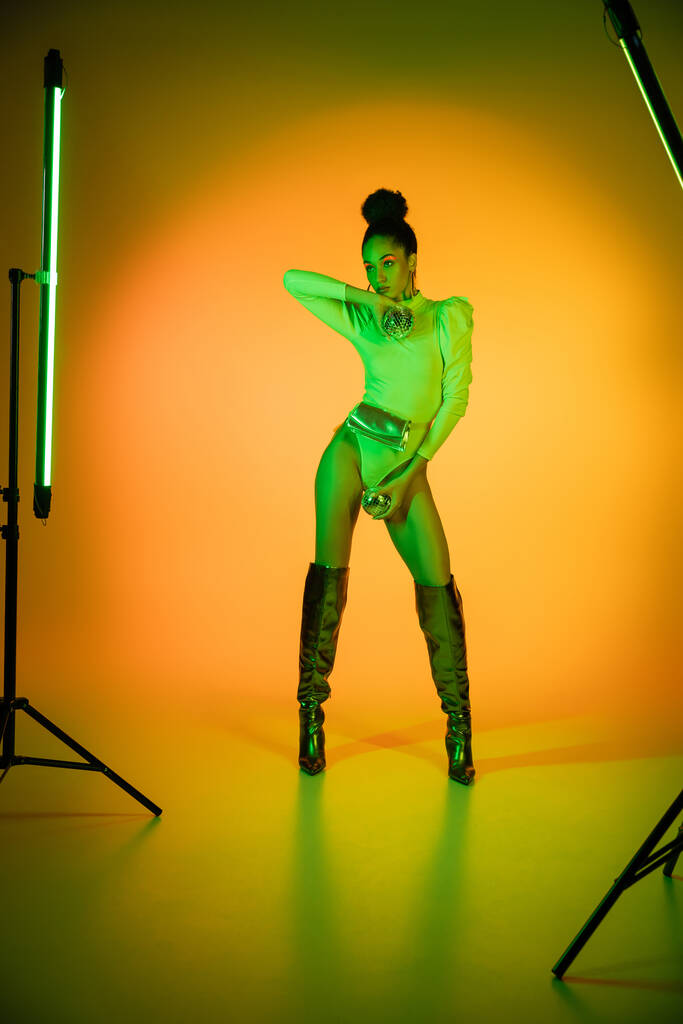 African american model in neon bodysuit and makeup holding disco balls near fluorescent lamps on orange background  - Photo, Image
