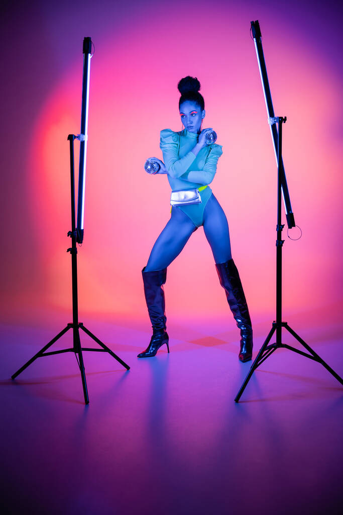 Trendy african american model in bodysuit holding disco balls near neon lamps on purple background - Photo, Image