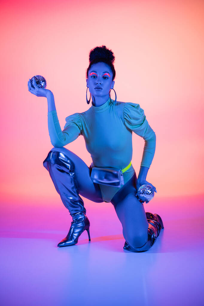 Módní africký americký model v bodysuit a kolena boty držení disco koule na růžovém a fialovém pozadí - Fotografie, Obrázek