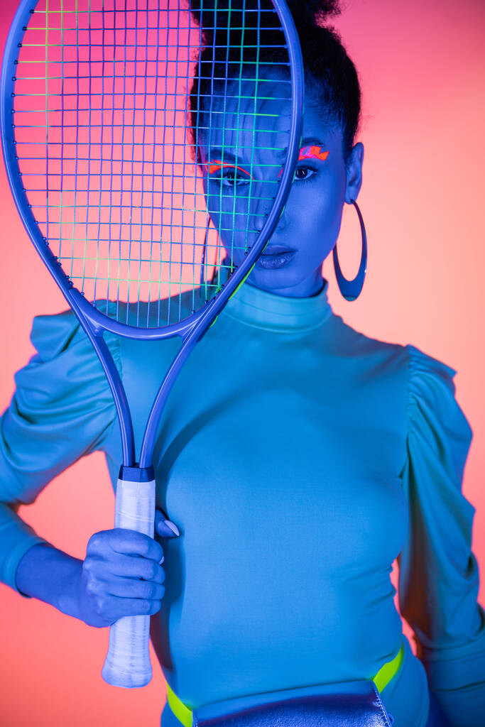 Portret van modieuze Afro-Amerikaanse vrouw met neon eyeliner houden tennis racket op roze achtergrond - Foto, afbeelding