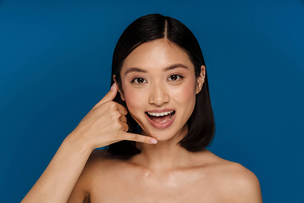 młody piękny szczęśliwy uśmiechnięty Azji kobieta z otwarte usta robi telefon gest patrząc w aparat, a stojąc nad odizolowanym niebieskim tle - Zdjęcie, obraz