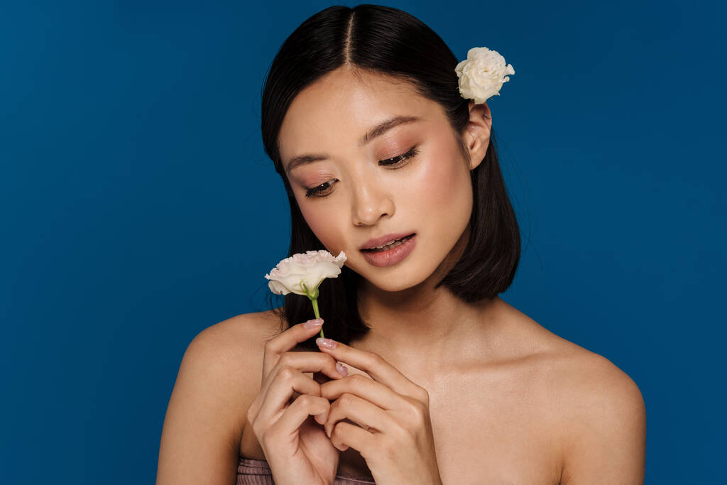 Mladá krásná asijská žena s otevřenými ústy s květinou na uchu drží bílou květinu a dívá se na to, zatímco stojí nad izolované modré pozadí - Fotografie, Obrázek