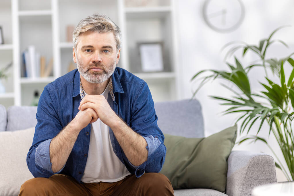 Retrato de un hombre de pelo gris guapo asustadizo con una camisa azul sentado en el sofá en casa y mirando seriamente a la cámara, cruzando sus brazos delante de él. - Foto, imagen