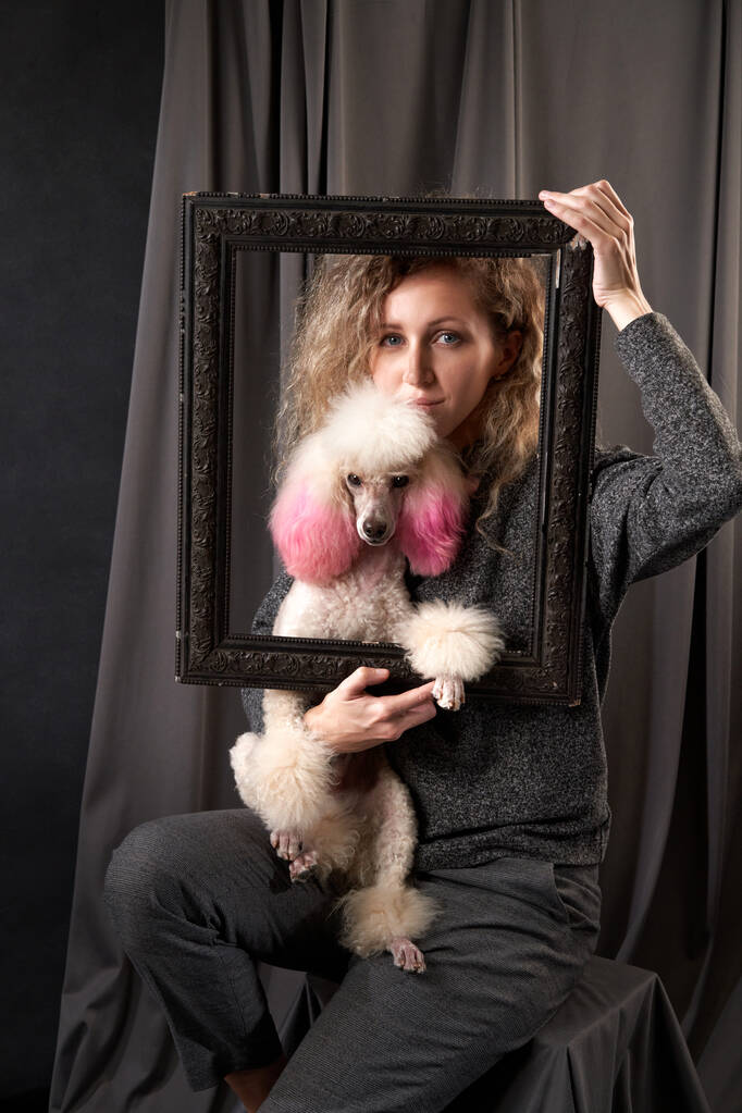 chica feliz con un perro en un estudio de fotografía en gris. Poodle blanco y mujer rizada - Foto, Imagen