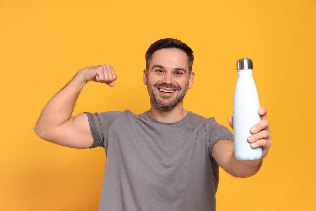 Hombre feliz sosteniendo la botella termo y mostrando el brazo sobre fondo naranja - Foto, imagen