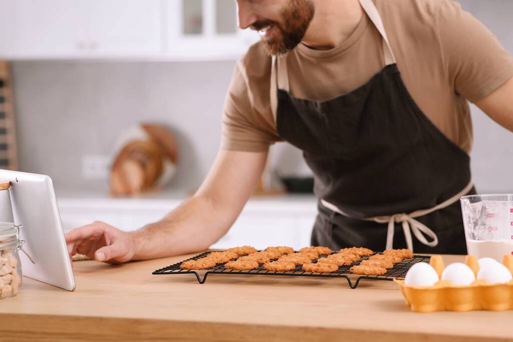 Mann mit frisch gebackenen Plätzchen schaut Online-Kochkurs via Tablet in Küche, Nahaufnahme - Foto, Bild