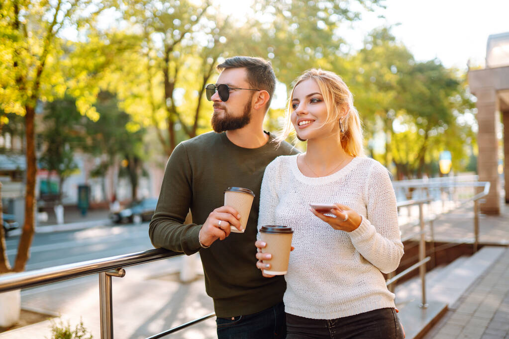 Jeune couple s'amuser à parler, boire du café et utiliser le téléphone tout en marchant dans la ville du matin. Relaxation, amour, jeune, mode de vie, selfie, blogging. - Photo, image