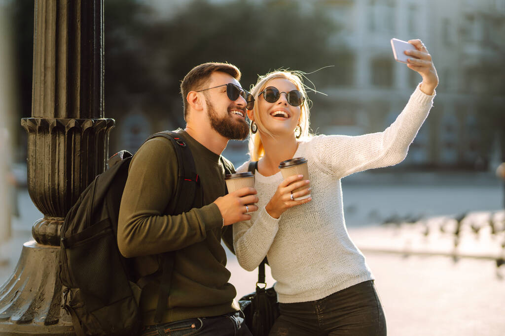 Νεαρό ζευγάρι διασκεδάζει μιλώντας, πίνοντας καφέ και χρησιμοποιώντας το τηλέφωνο, ενώ περπατάτε στην πόλη το πρωί. Χαλάρωση, νιάτα, αγάπη, τρόπος ζωής, selfie, blogging. - Φωτογραφία, εικόνα