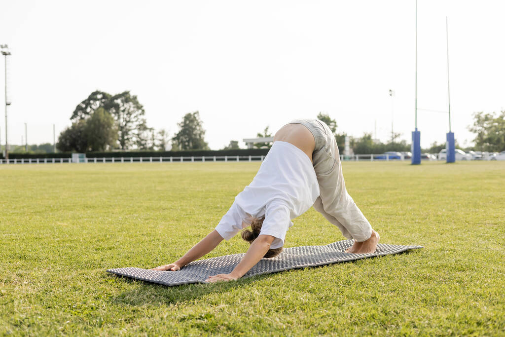 homme pieds nus en t-shirt blanc et pantalon en coton pratiquant le yoga en pose de dauphin sur la pelouse verte à l'extérieur - Photo, image