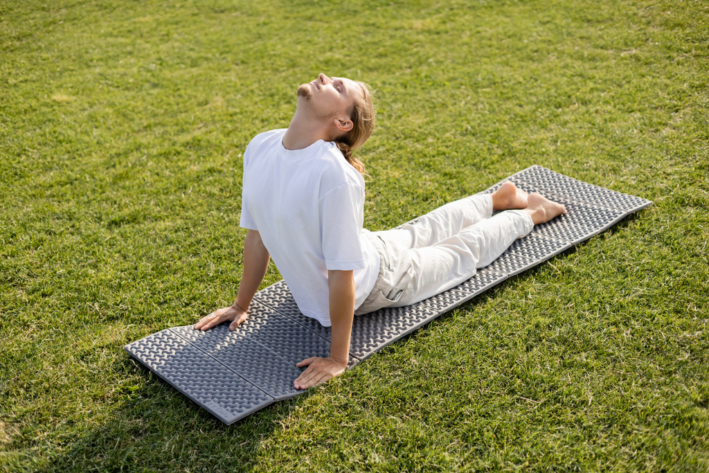 Beyaz tişörtlü ve keten pantolonlu çıplak ayaklı bir adam yeşil çimlerde kobra pozisyonunda yoga yapıyor.  - Fotoğraf, Görsel